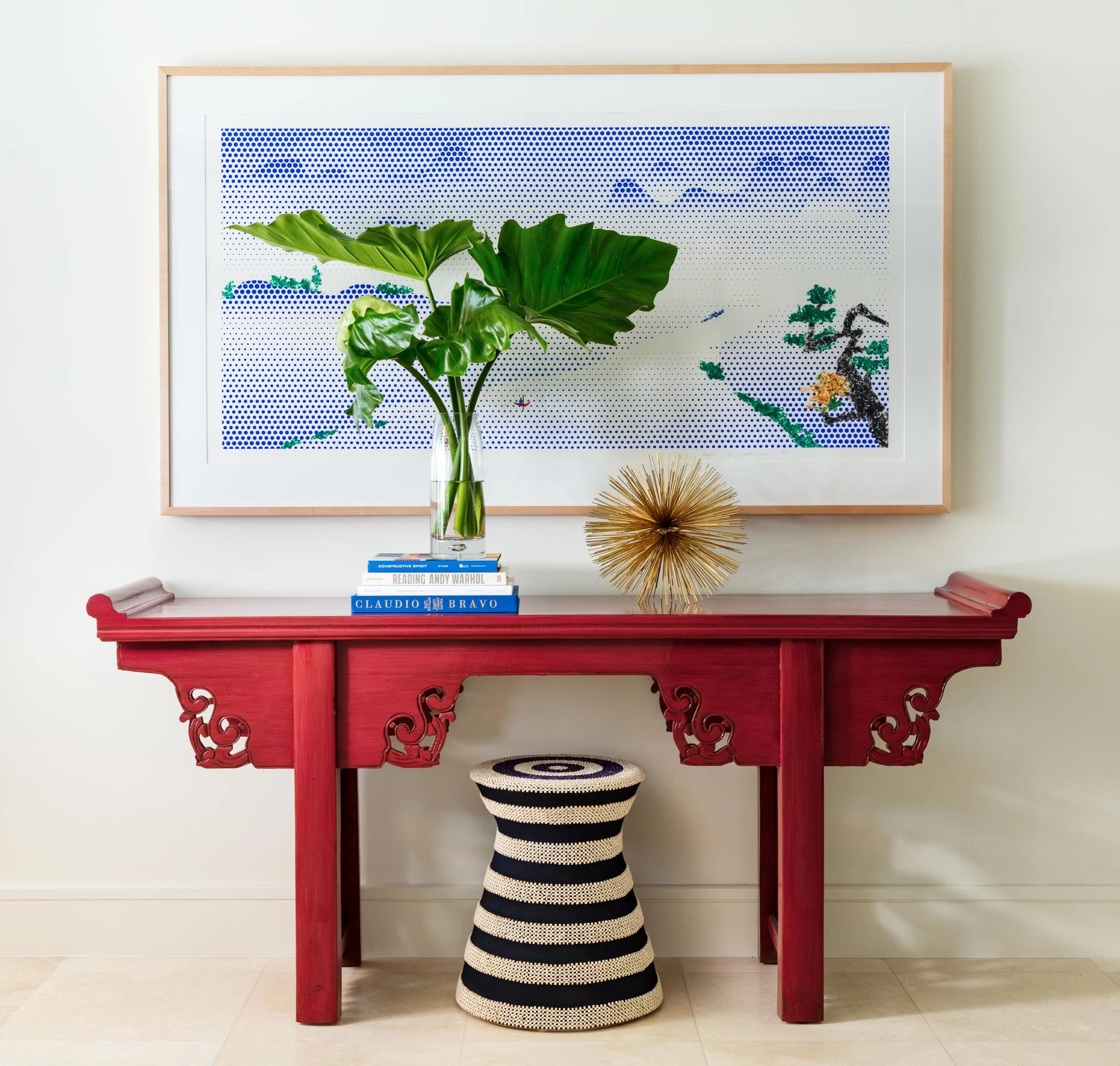 Step-by-step guide. Gorgeous red console table in a bright hallway. There is a blue landscape art piece hanging above the table. On the table, there are books, a plant, and a gold decorative ball that looks like a sea urchin. Under the table sits a black and white striped decorative foot stool.